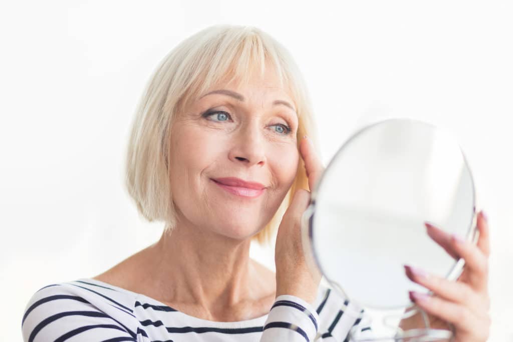 Senior woman touching her soft face skin, looking in mirror happy at her liquid facelift results.