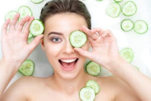 overhead headshot of woman laying on solid white surface holding a sliced cucumber over her eye smiling playfully