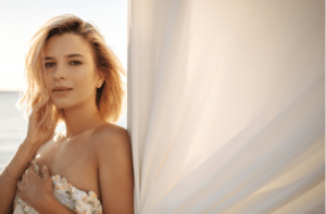Short haired blonde looks tenderly at camera dressed in white sundress with bare shoulders.